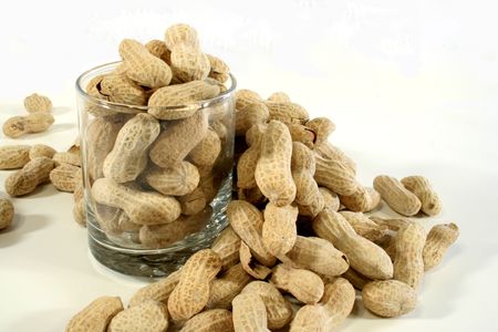 Peanuts in a clear glass with more peanuts spilled around the glass on a white background.