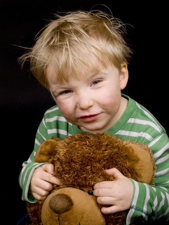 Smiling little boy with brown teddy-bear on black background. Boy have blue eyes, blond hair and a bit of dirt on his faceの写真素材