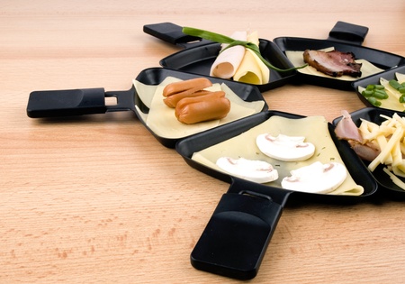 Raclette pans with edam cheese and ham, bacon, mushroom, spring onion, coctail sausages on wooden tableの写真素材