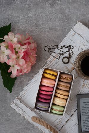 Colorful macarons into a box on the white wooden table background. Macaroons close up. Cup of tea coffee. Pink flowers. Flat lay top view.の写真素材