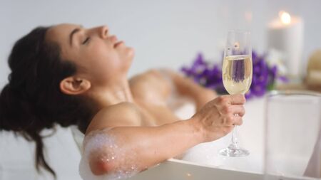 Sexy girl relaxing with glass of wine in bathtub. Brunette woman holding champagne at luxury bath. Closeup relaxed woman washing legs with foam at home bathroom.の写真素材