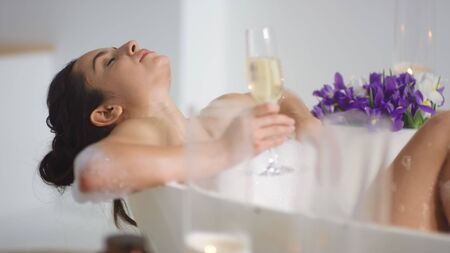 Close up cute girl resting in modern bathtub. Relaxed girl lying in bath with glass of wine. Side...