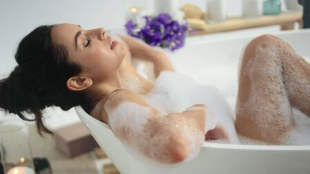 Closeup relaxed lady lying in bathtub with foam. Romantic woman sleeping in bath at luxury home. Side view of sensual girl enjoying foam bath in slow motion.の写真素材