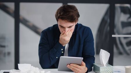 Focused business man reading tablet computer in office. Ill man getting flu in office. Sick businessman sneezing at work place in slow motion. Unhealthy guy using wipe for nose treatment.の写真素材