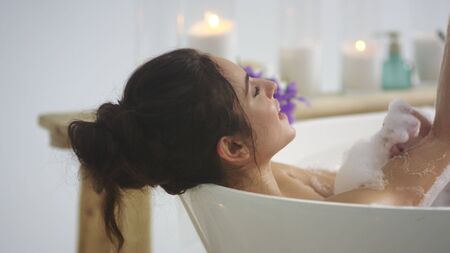Close up relaxed woman touching skin with foam in bath. Side view of sexy girl taking bathtub at home. Romantic brunette woman relaxing bath in slow motion.の写真素材