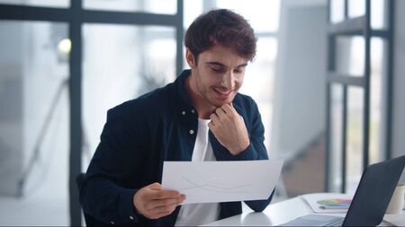 Smiling business man reading graphs research in home office. Cheerful professional comparing results in papers and laptop. Close up happy businessman looking papers in modern office.の写真素材