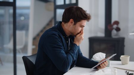 Sick business man getting flu in office. Ill man sneezing near laptop indoor. Young guy feeling unhealthy. Ill businessman using wipe for nose.の写真素材