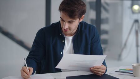 Close up happy businessman working with documents in modern office. Smiling guy making notes on financial diagram in slow motion. Cheerful man enjoying good results of project at workplace.の写真素材