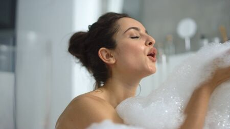 Closeup hot woman blowing foam at bathtub in slow motion. Side view of sexy girl playing with bubbles in bathroom at home. Sensual woman having fun with foam in bath.の写真素材