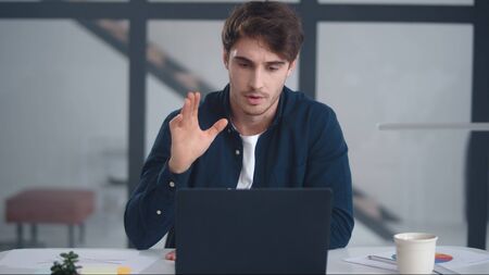 Serious business man talking on video conference in office. Focused man explaining project with gestures at video chat online on laptop computer. Male professional having video call on notebook indoorの写真素材