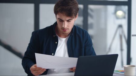 Focused businessman reading document carefully. Serious professional comparing results in papers and laptop in slow motion. Closeup thoughtful man looking papers in modern office.の写真素材