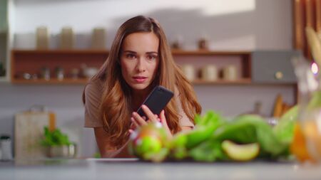 Smiling food blogger taking photo on mobile phone of fresh vegetables on kitchen. Woman using smartphone at home. Cheerful girl standing on domestic kitchen in slow motionの写真素材
