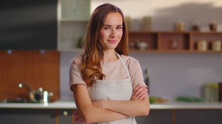 Cheerful woman with crossed hands standing on kitchen. Smiling housewife looking at camera on modern kitchen in slow motion. Portrait of young woman posing in cooker apron at homeの写真素材