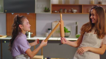 Happy daughter and mother fighting with rolling pins on domestic kitchen. Joyful woman and girl...