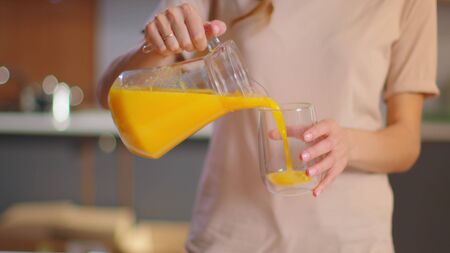 Close up of woman pouring orange juice into the glass from jar on kitchen in morning. Girl holding glass of fruit juice at home. Smiling lady drinking orange juice on modern kitchen in slow motionの写真素材