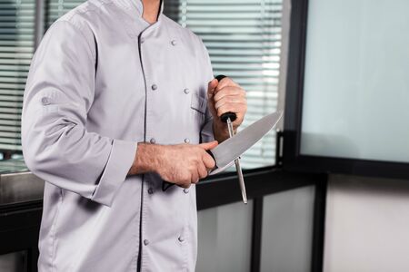 Chef man hands with knife at kitchen restaurant. Closeup chef hands with knife. Male professional in uniform sharpen knife at commercial kitchen.の写真素材