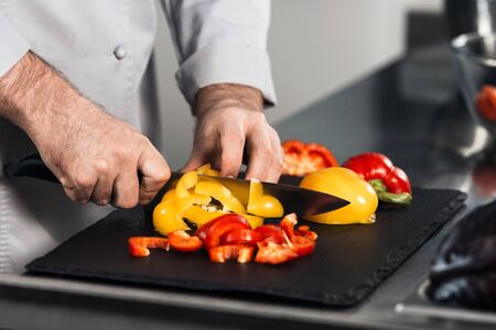 Chef hands cook food at kitchen restaurant. Closeup chef hands slice yellow and red pepper at kitchen restaurant. Male cooker is cutting vegetables with knife for healthy food.の写真素材