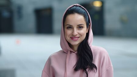 Closeup of beautiful woman looking camera outdoor. Fitness woman posing to camera in sport clothes. Portrait of smiling girl in hoodie on city street.の写真素材