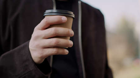 Closeup attractive man hand holding paper cup outdoors. Hipster guy hand bringing takeaway coffee to...