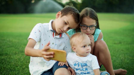 Focused kids sitting on green grass in summer meadow. Cute boy taking selfie photo on cellphone with sister and brother in green park. Adorable children looking smartphone screen outdoorsの写真素材