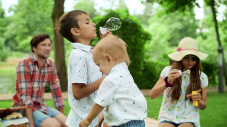 Cheerful children blowing bubbles outdoors. Smiling parents spending time with kids in park. Young family playing with soap bubbles outside. Positive parents relaxing with siblings in forestの写真素材