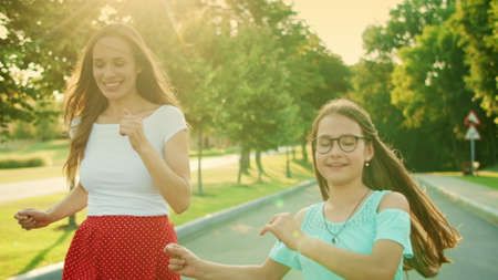 Portrait of happy girl and woman dancing in park. Joyful mother and daughter gesturing with hands during dance. Female parent and child making dance movements on street. Family having fun outdoorsの写真素材