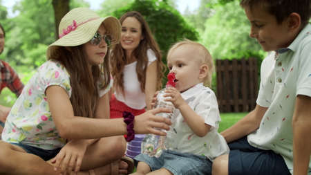 Happy family sitting on blanket in park. Cute sister helping small brother with water bottle. Adorable toddler drinking water from bottle outdoors. Joyful parents spending weekend with kidsの写真素材