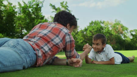 Focused father and son practising arm wrestling in meadow. Happy boy winning competition in armwrestling in park. Smiling boy celebrating success. Positive family playing together outdoorsの写真素材