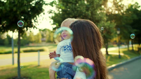 Portrait of happy mother holding son on hands in summer park. Closeup smiling woman and toddler playing with soap bubbles outside. Joyful boy and female parent spending time togetherの写真素材