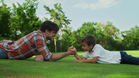 Side view cheerful boy and man armwrestling on green grass in meadow. Closeup father and son practising arm wrestling together outdoors. Happy boy winning competition in armwrestling at summer parkの写真素材