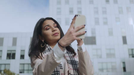 Cheerful businesswoman taking selfie photo on smartphone at city street. Smiling indian girl using...