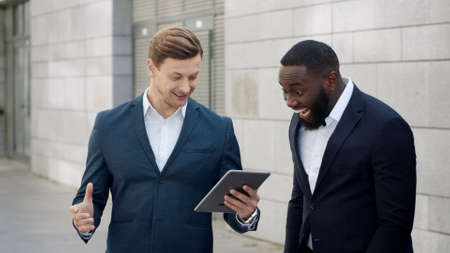 Surprised businessmen reading good news on digital tablet at street. Smiling male professionals celebrating victory outside. Excited business men hugging each other in slow motionの写真素材
