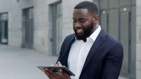 Portrait of happy afro business man typing message on digital tablet at modern street. Smiling male worker in formal wear using pad outdoors. Successful businessman working on touchpad in slow motionの写真素材