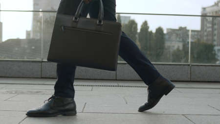 Close up unrecognizable businessman legs walking on urban street. Business man with briefcase going for job in slow motion. Male worker taking walk around cityの写真素材