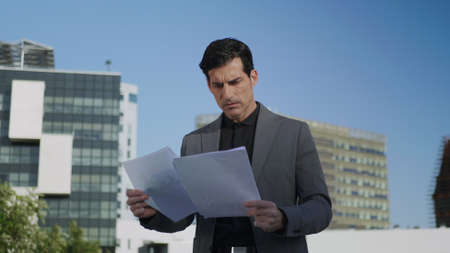Portrait of serious businessman looking papers on city street. Worried manager reading documents in...