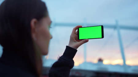 Portrait of attractive girl taking picture on cellphone with green screen. Closeup focused woman making video on smartphone on city street. Girl using mobile phone with chromakey for selfie photoの写真素材