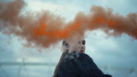 Closeup attractive girl waving hand at camera with smoke bomb. Young woman posing at camera. Hipster girl using smoke grenade at street riot. Portrait of trendy girl looking at camera outdoorsの写真素材