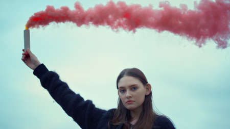 Focused girl holding colorful smoke bomb in hand at street protest. Closeup attractive woman using...