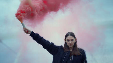 Portrait of young woman standing on street with smoke bomb in hand. Ambitious girl holding active smoke grenade at protest. Closeup serious activist looking at camera on urban streetの写真素材