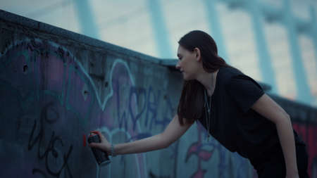 Attractive girl painting graffiti on urban street. Young woman using spray bottle for graffiti. Female artist drawing infinity sign with red color. Girl drawing graffiti with spray paint on wallの写真素材