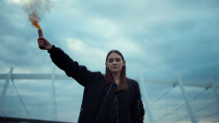 Portrait of ambitious girl protesting on street with smoke bomb in hand. Serious woman holding sparkling smoke grenade at protest. Closeup young activist posing at camera with smoke flareの写真素材
