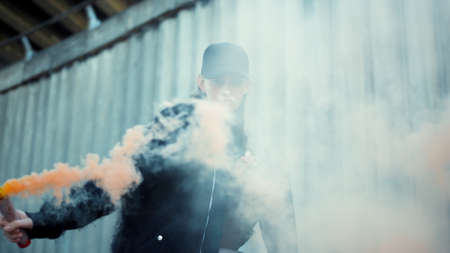 Stylish woman posing at camera with smoke bomb in hand. Riot girl waving smoke grenade on city street. Female activist protesting with colorful smoke outdoorsの写真素材