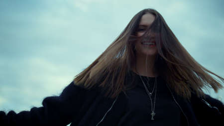 Positive girl showing peace sign at camera. Cheerful woman showing v sign. Hipster with raised hands turning around on street. Attractive woman waving head with hair. Female model smiling at cameraの写真素材