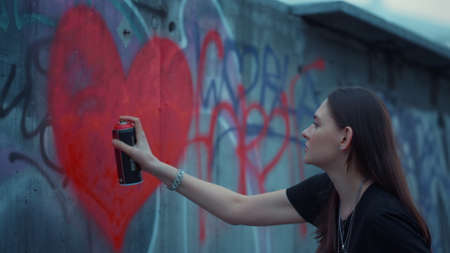 Focused girl drawing graffiti on wall. Focused woman painting heart with spray bottle. Portrait of young woman using red color for graffiti. Female artist making street art with aerosol paintの写真素材