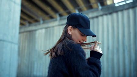 Smiling girl flirting with camera outdoors. Portrait of stylish woman playing with hair at windy weather. Young lady taking off cap from head. Closeup hipster posing on city streetの写真素材