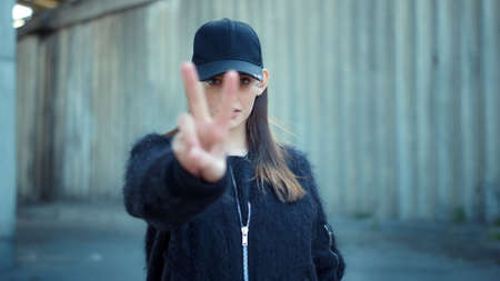 Closeup stylish woman show v sign on urban street. Serious girl showing peace sign outdoors. Smiling girl activist showing fuck you sign at protest. Positive hipster doing offensive gesture at cameraの写真素材