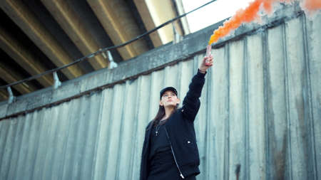 Female activist protesting on street with smoke bomb in hand. Beautiful woman holding colorful smoke flare. Serious girl turning around on street. Riot girl posing at camera outdoorsの写真素材