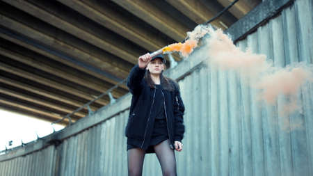 Attractive girl holding smoke bomb on urban street. Serious woman protesting with colorful smoke grenade. Hipster girl posing at camera with smoke flare at street riot. Female activist portraitの写真素材