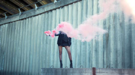 Stylish girl standing on street with smoke bomb. Young woman holding smoke grenade. Hipster wearing protective mask during protest. Portrait of activist posing on urban street in colorful smokeの写真素材