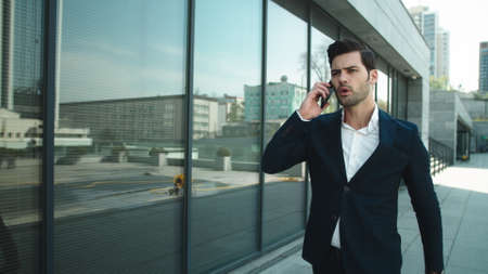 Close up serious man walking in slow motion in suit with phone outdoors. Confused businessman receiving bad news at street. Annoyed business man talking smartphone at modern street.の写真素材